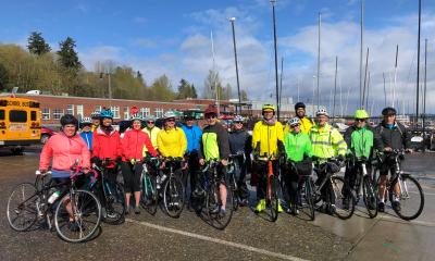 A group of riders in colorful rain jackets in yellow, green, and pink, stand in front of Cascade Bicycle Club and Sandpoint Sail's sailboats.